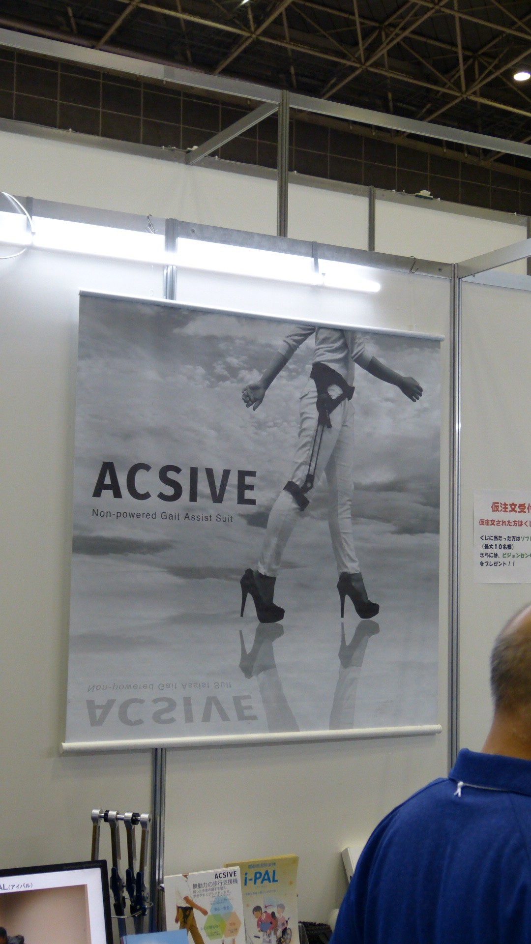 a poster depicts a woman's body walking in high heeled shoes with a brace around her waist and one of her legs, below the knee.  Text reads Acsive non-powered gait assist suit